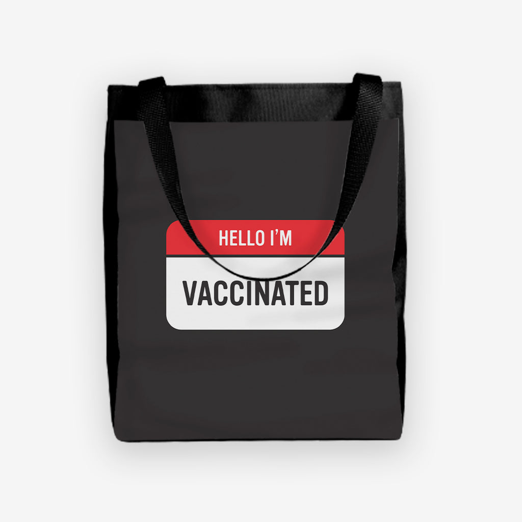A black day tote with a red and white label at the center. The red part of the label reads “Hello I’m” in white text. The white part of the label reads “Vaccinated” in black text.