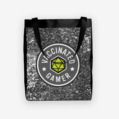 A day tote with grey, white, and black splattered paint behind a circular logo. The logo is white and grey with a green 20 sided dice in the middle. The white text around the dice reads “Vaccinated Gamer.” “Vaccinated” is separated from “Gamer” by two white stars.