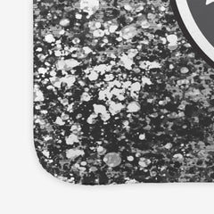 A close-up of a large gaming mousepad with grey, white, and black splattered paint behind a circular logo.
