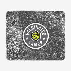 A large gaming mousepad with grey, white, and black splattered paint behind a circular logo. The logo is white and grey with a green 20 sided dice in the middle. The white text around the dice reads “Vaccinated Gamer.” “Vaccinated” is separated from “Gamer” by two white stars.