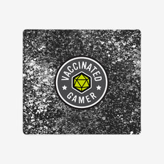 A extra large gaming mousepad with grey, white, and black splattered paint behind a circular logo. The logo is white and grey with a green 20 sided dice in the middle. The white text around the dice reads “Vaccinated Gamer.” “Vaccinated” is separated from “Gamer” by two white stars.