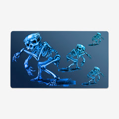 Playmat of Creeping Skelly by Why Try Designs. One large skeleton faces to the left, it is hunched over. Three other smaller skeletons face to the right and are also crouched over. All four skeletons are blue and white on a blue background.